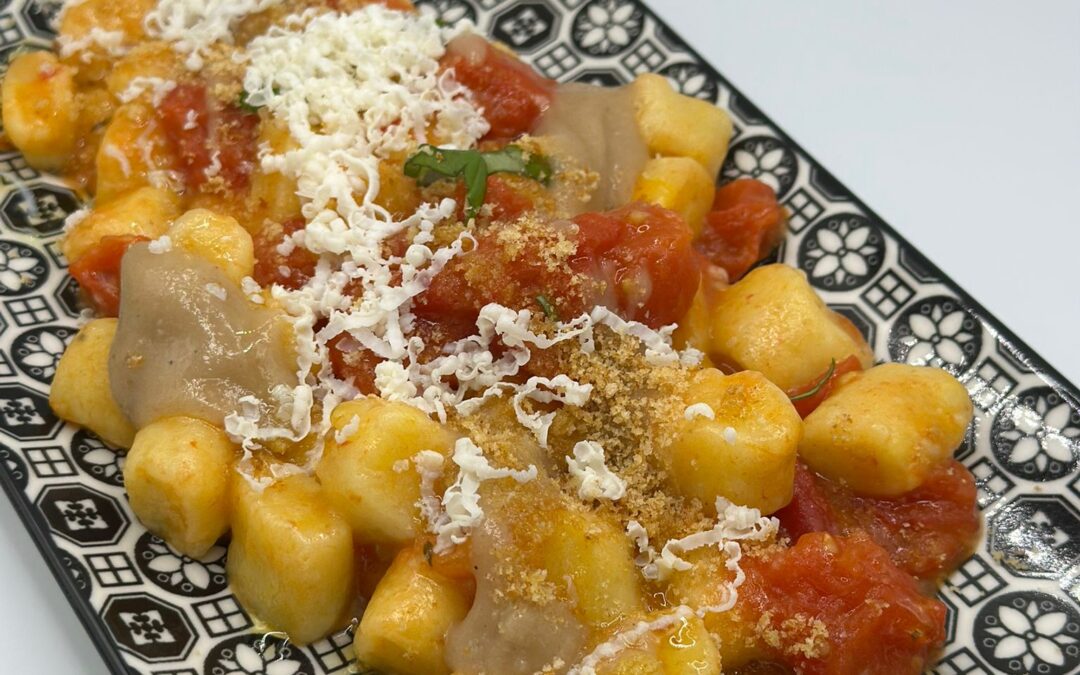 Un piatto rettangolare decorato con gnocchi al datterino conditi con salsa di pomodoro, formaggio grattugiato, pangrattato ed erbe aromatiche fresche, serviti su uno sfondo bianco.