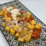 Un piatto rettangolare decorato con gnocchi al datterino conditi con salsa di pomodoro, formaggio grattugiato, pangrattato ed erbe aromatiche fresche, serviti su uno sfondo bianco.